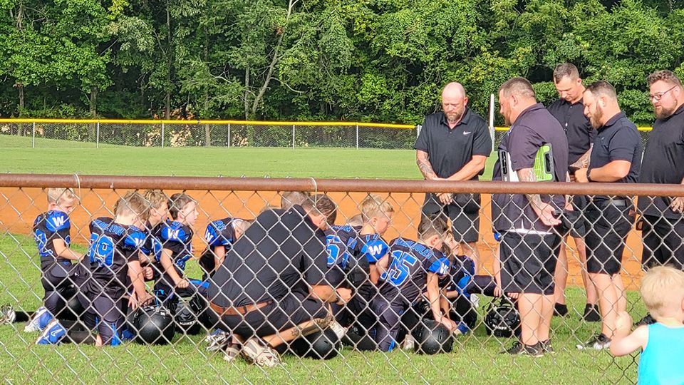 A group of football players are kneeling on the field with their coaches.