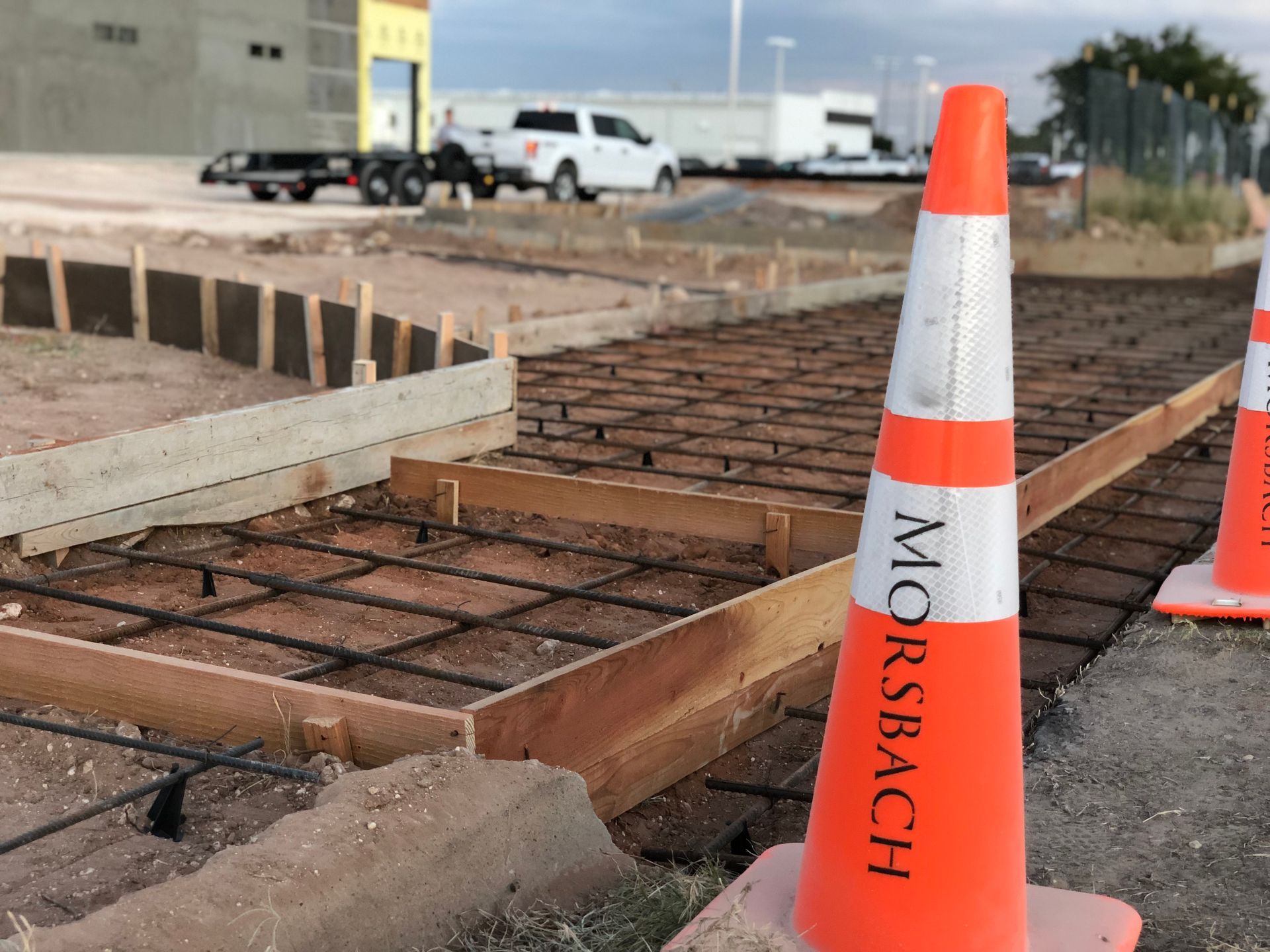 Construction site with orange cones, rebar, and wooden forms. A truck and trailer are visible in the background.