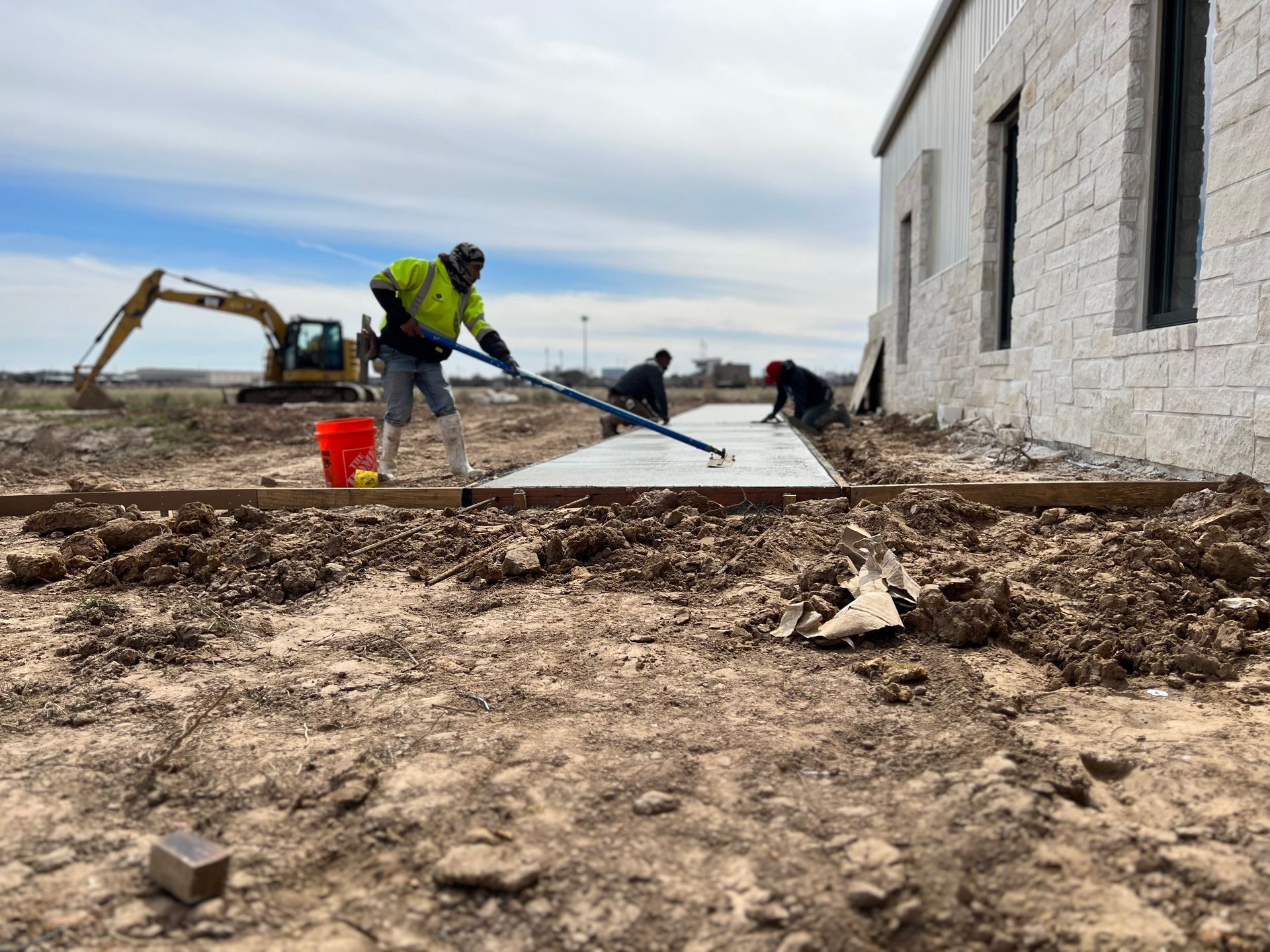 Construction workers smoothing wet concrete next to a building. An excavator sits in the background.