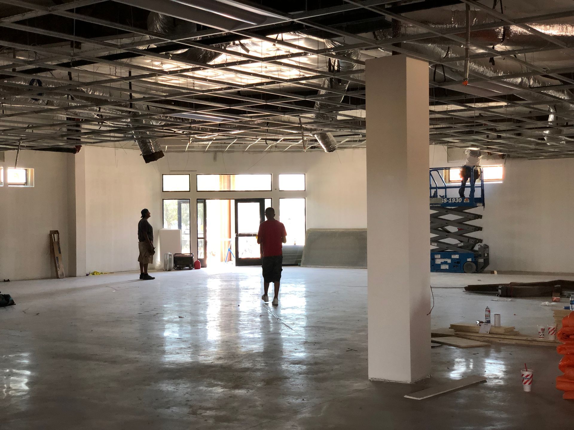 Interior construction site: Two workers and a lift in a large, open space with unfinished ceiling and concrete floor.