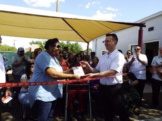Man in white shirt hands document to woman in blue shirt, cutting red ribbon at outdoor ceremony.