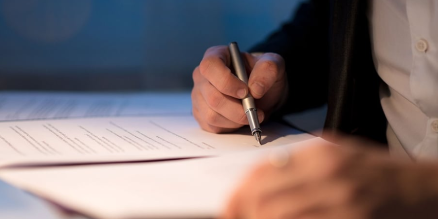 Person signing a document with a pen at a desk under soft light