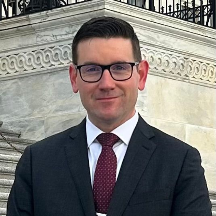 Man in glasses and suit standing outdoors in front of stone steps and ornate building railing