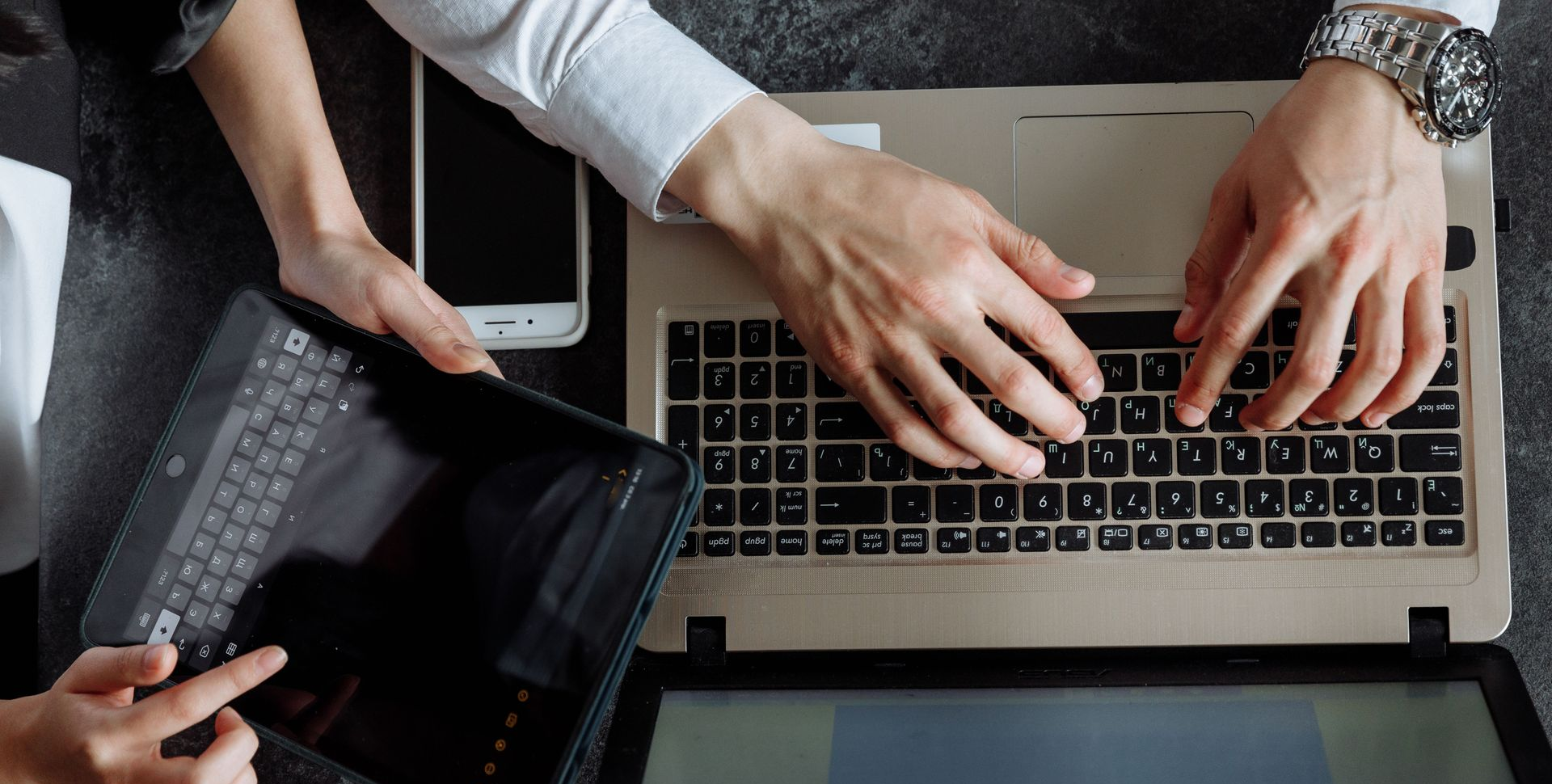 Hands typing on a laptop beside a tablet and smartphone on a desk