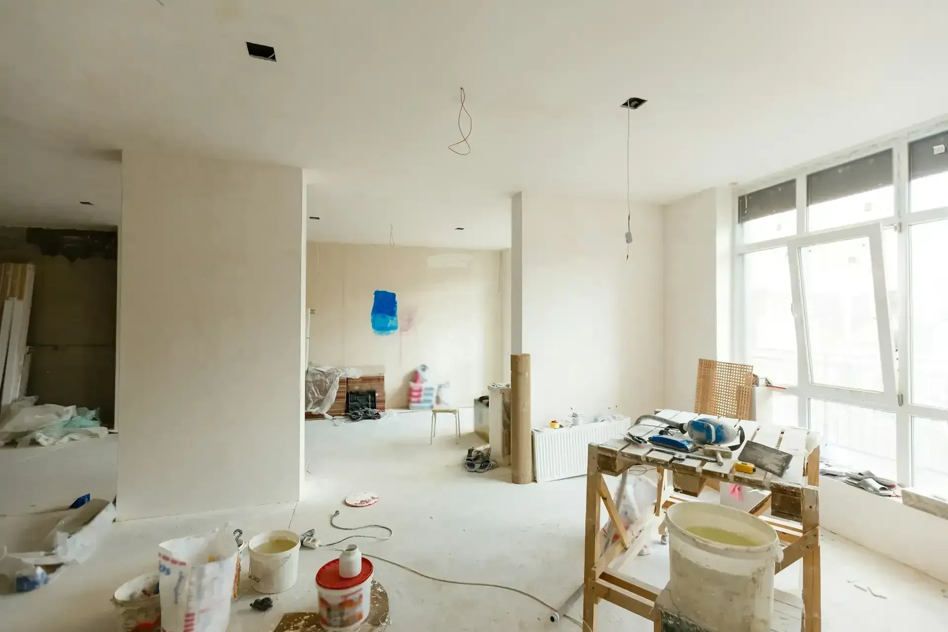 Bright, unfinished apartment interior with scattered paint supplies, boxes, and large windows.