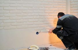 Worker spray-painting a beige brick wall near an outlet with orange light reflecting on the right side
