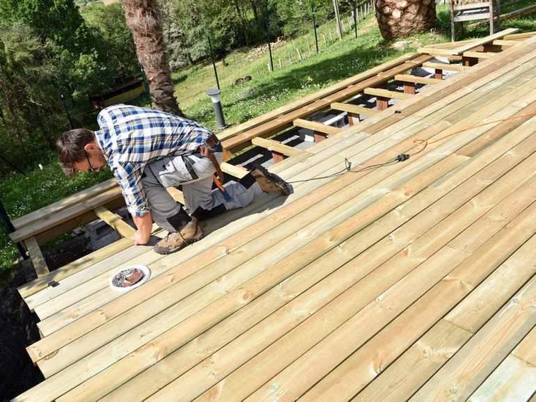 Person kneeling on a wooden deck under construction, placing boards with a tool outdoors.
