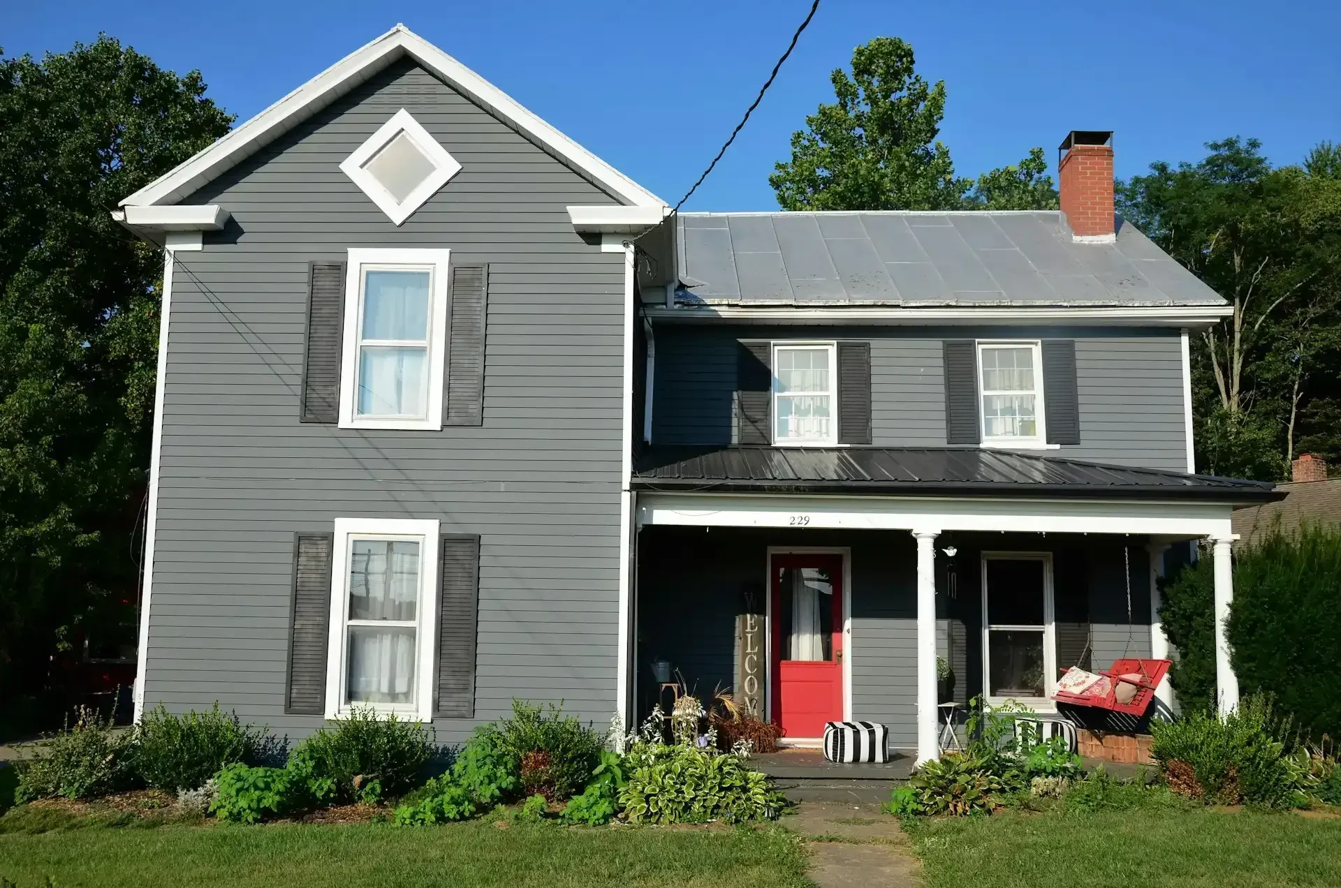 Gray two-story house with white trim, red door, and front porch in a sunny yard