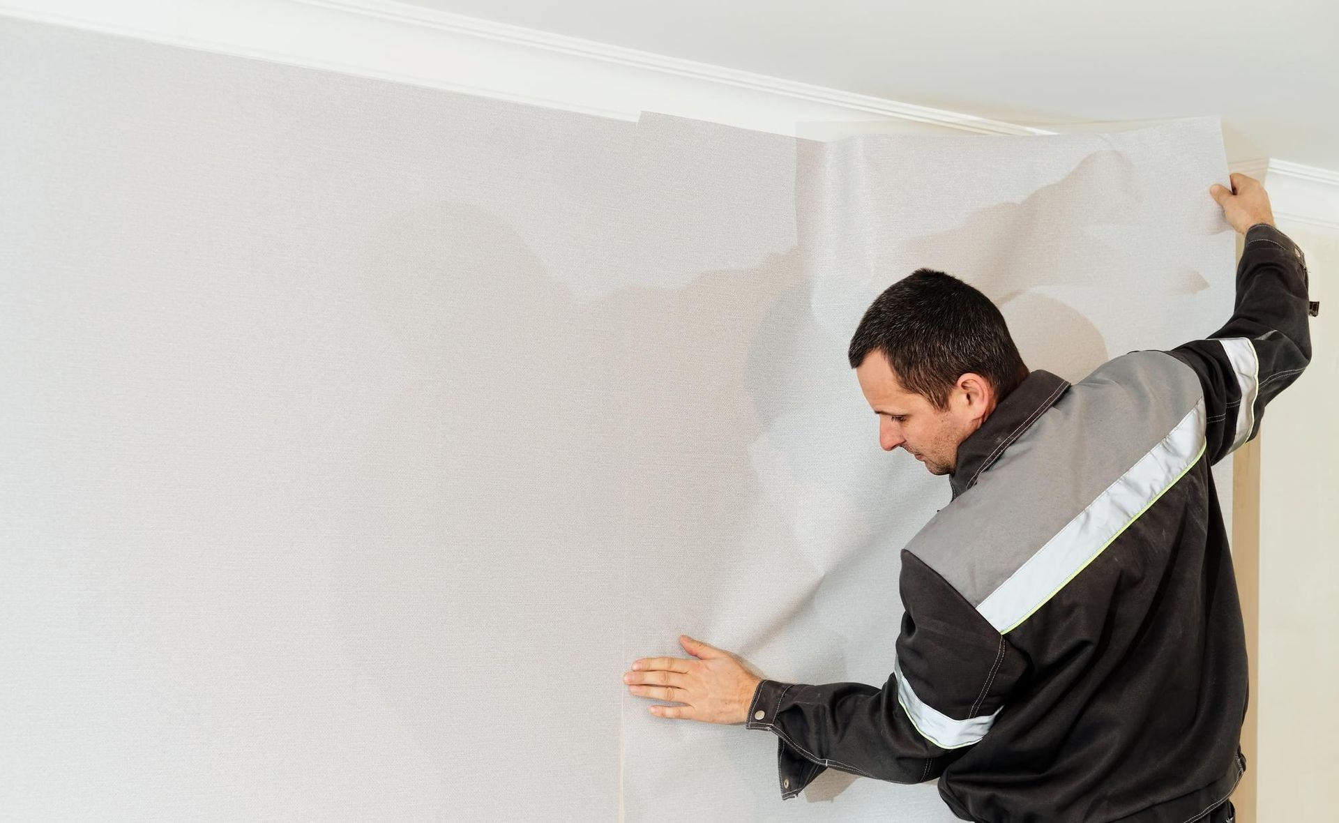 Man hanging wallpaper, smoothing a light gray wall with a tool in a bright room
