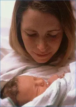 Woman gently looking at a sleeping newborn wrapped in a white blanket
