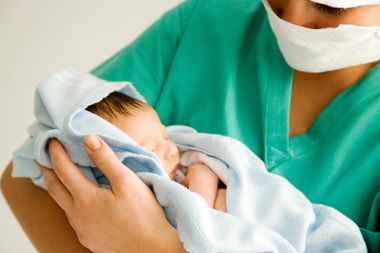 Newborn wrapped in a white blanket, held by a person in green medical scrubs and a face mask