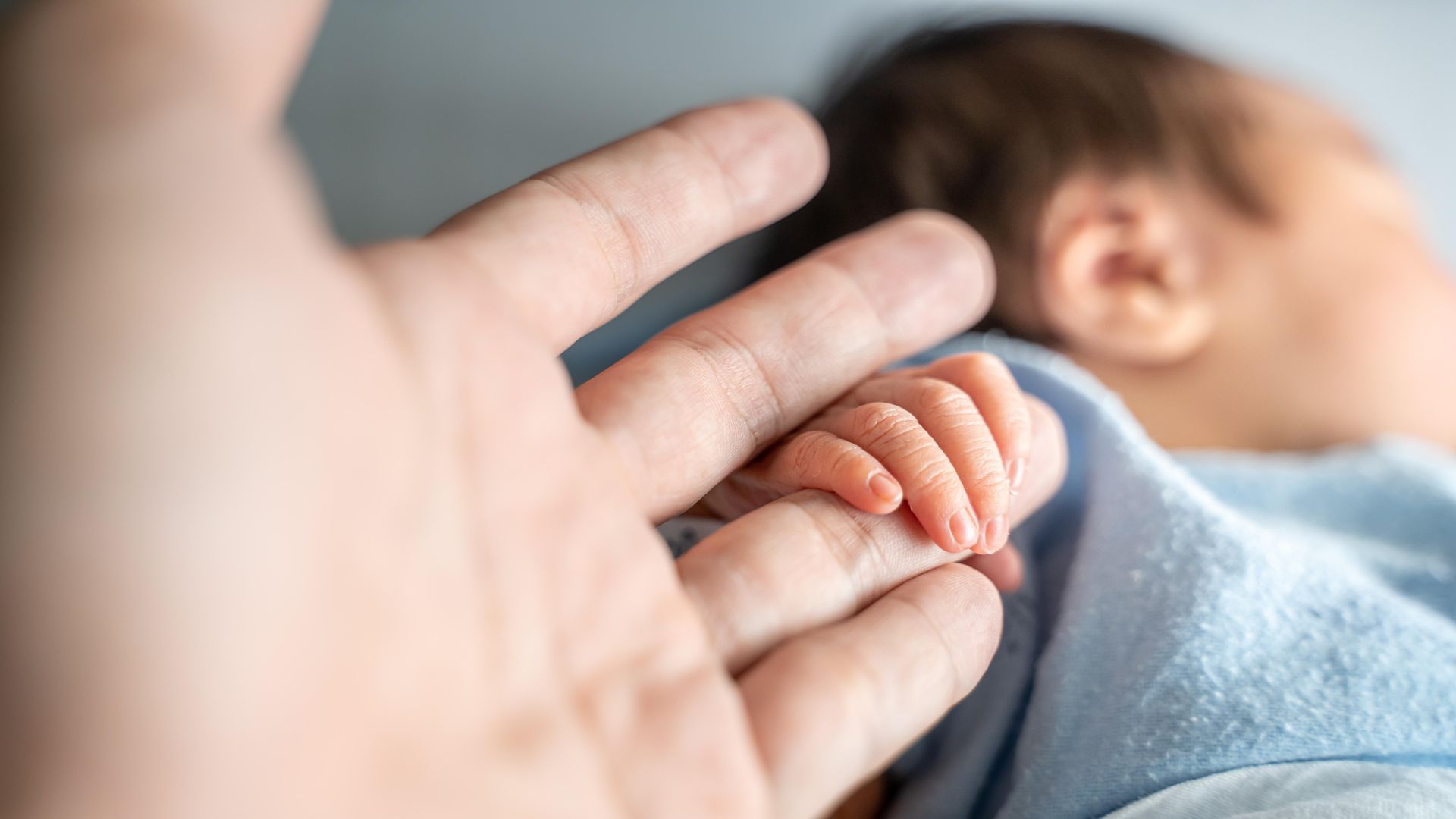 Adult hand holding a babyโs hand, with the baby lying on a blanket in the background