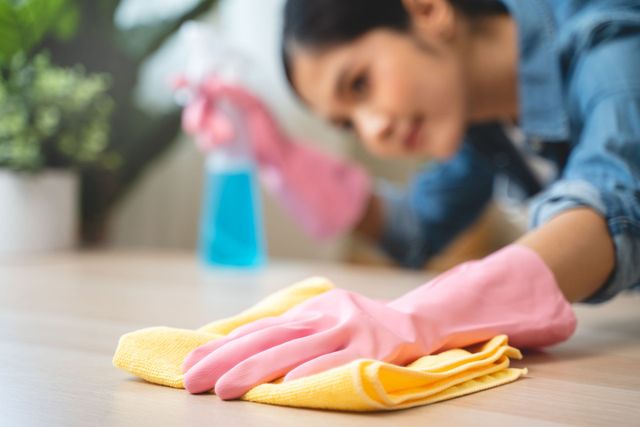 Person wearing pink gloves wiping a table with a yellow cloth and spray bottle in the background