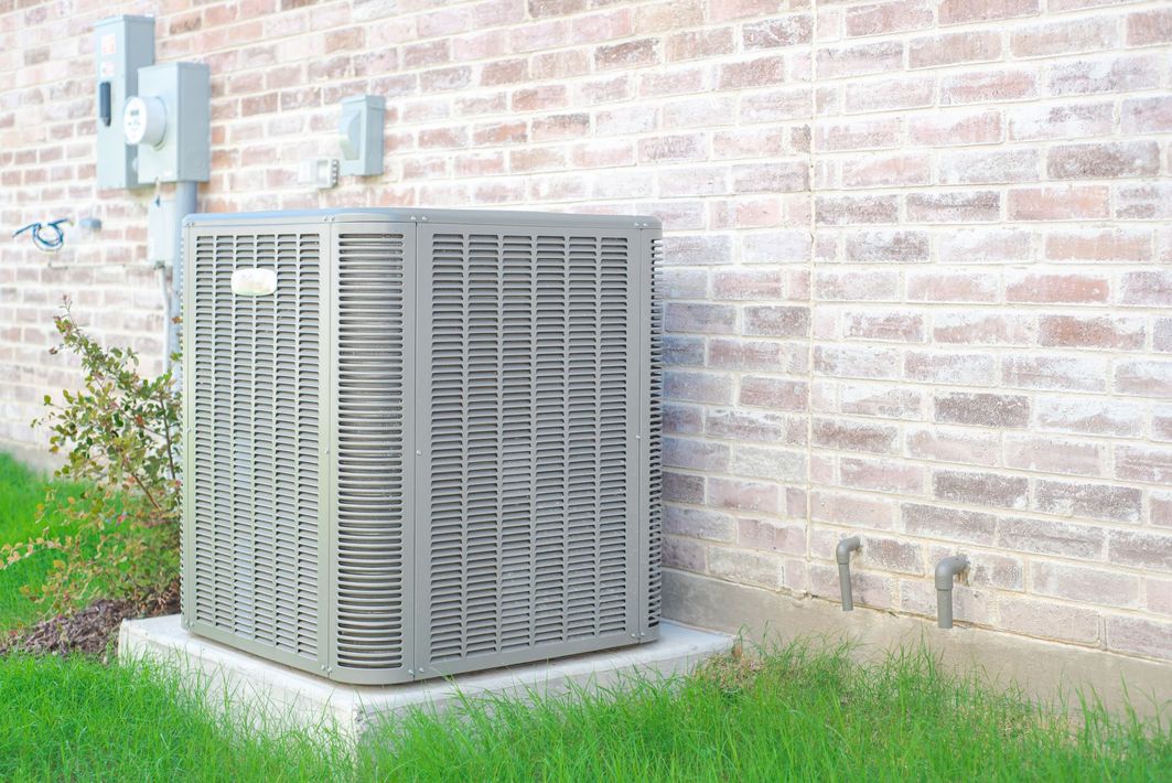Outdoor HVAC unit beside a brick wall, surrounded by grass and small plants.