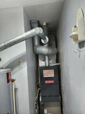 Furnace in a cramped closet with insulated ducts and a utility pipe along the wall.