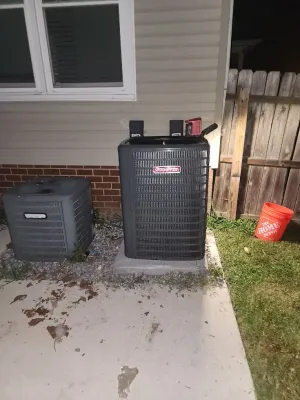 Two outdoor HVAC units beside a house wall and window, with a red bucket near a wooden fence.