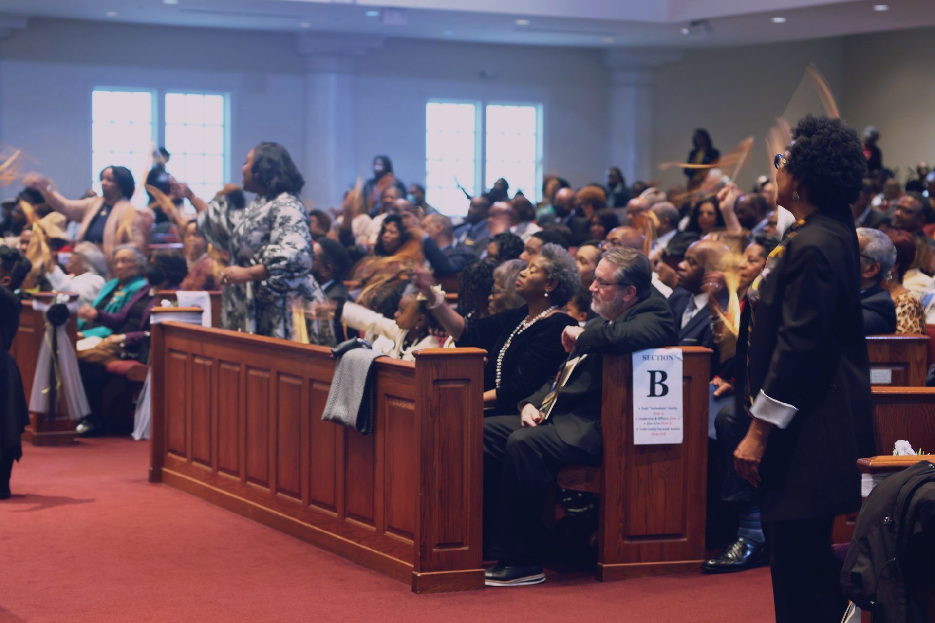 A church congregation gathers, with many people standing or raising their hands in a brightly lit auditorium.