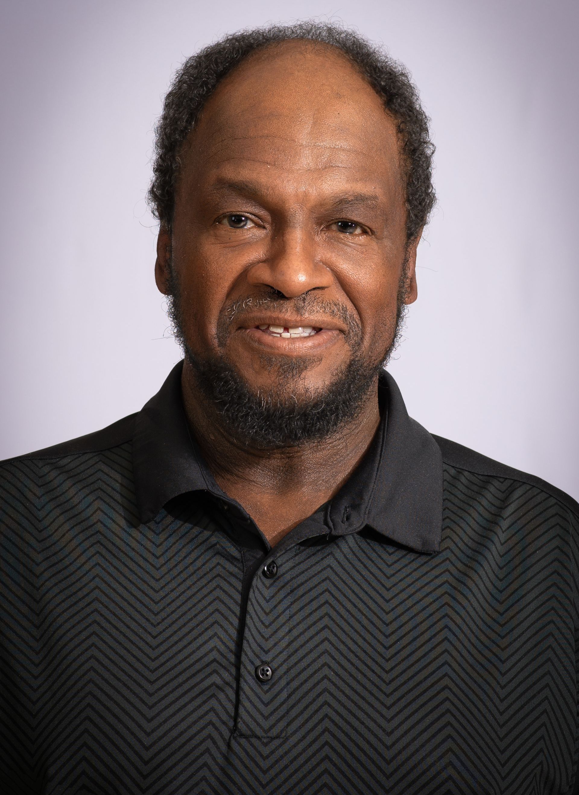 Headshot of a man with a beard, wearing a black textured polo shirt against a light grey background.