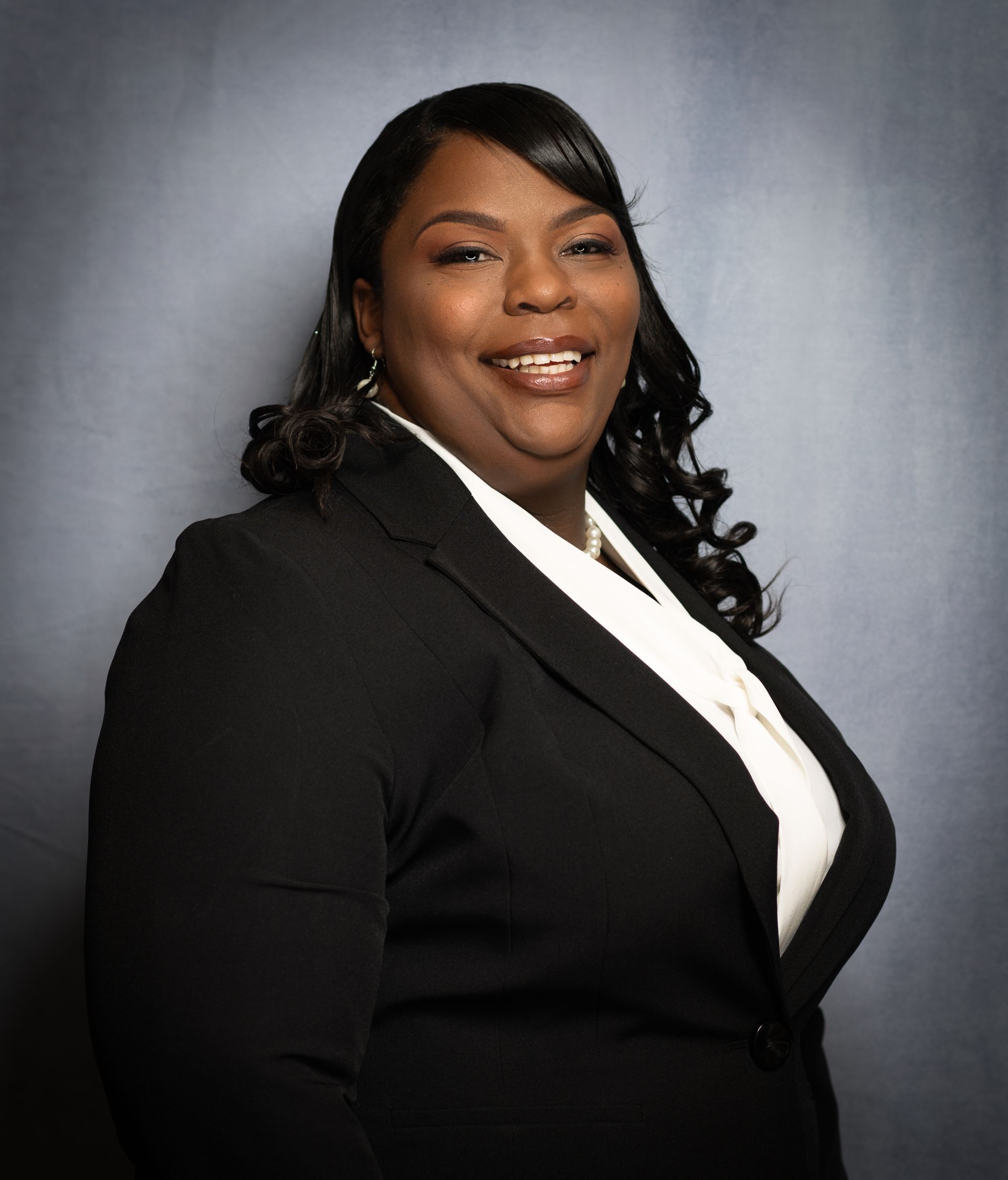 A smiling person posing in a black blazer and white top against a mottled grey background.