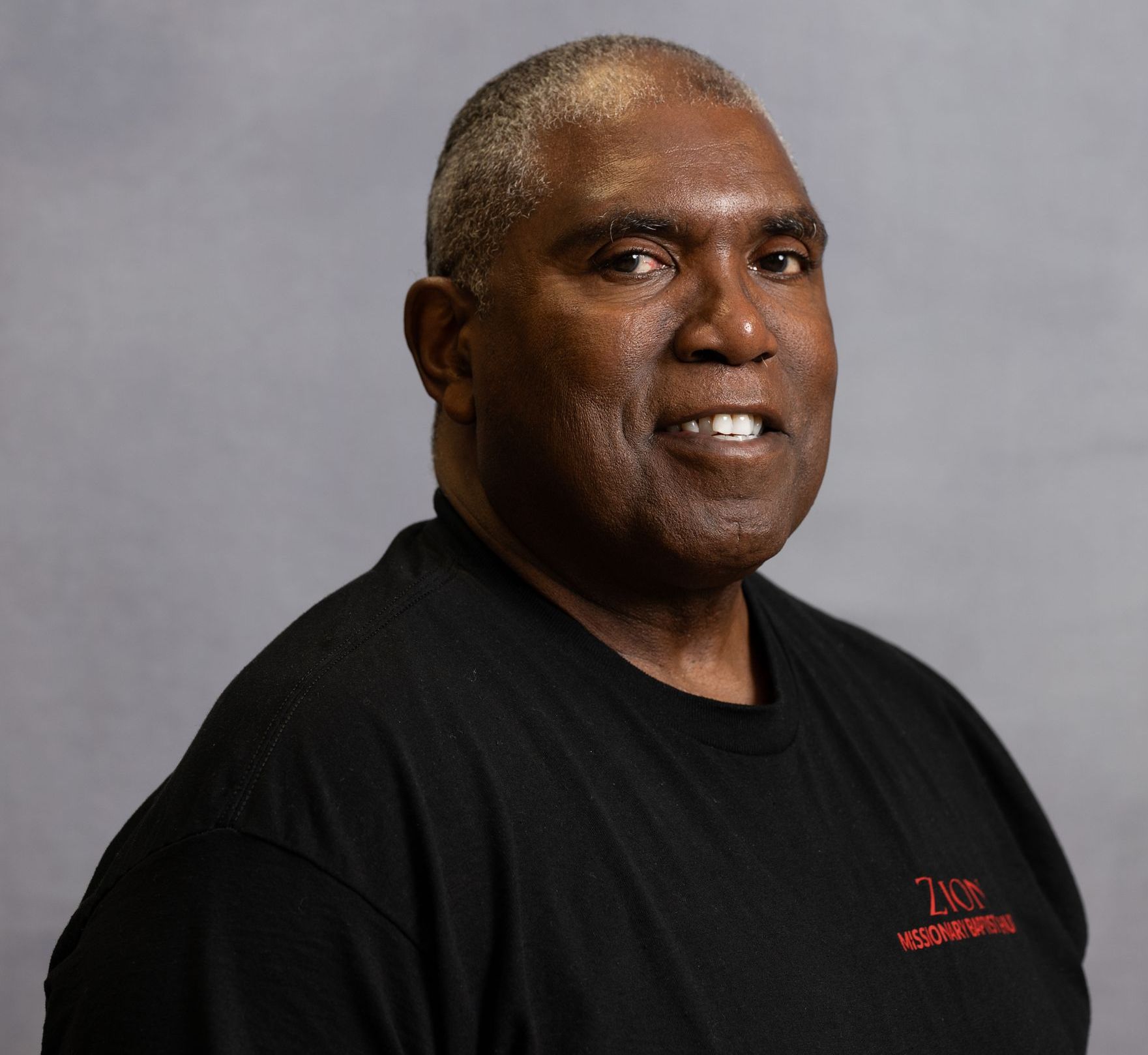 A person smiling against a gray background, wearing a black shirt with red text on the chest.