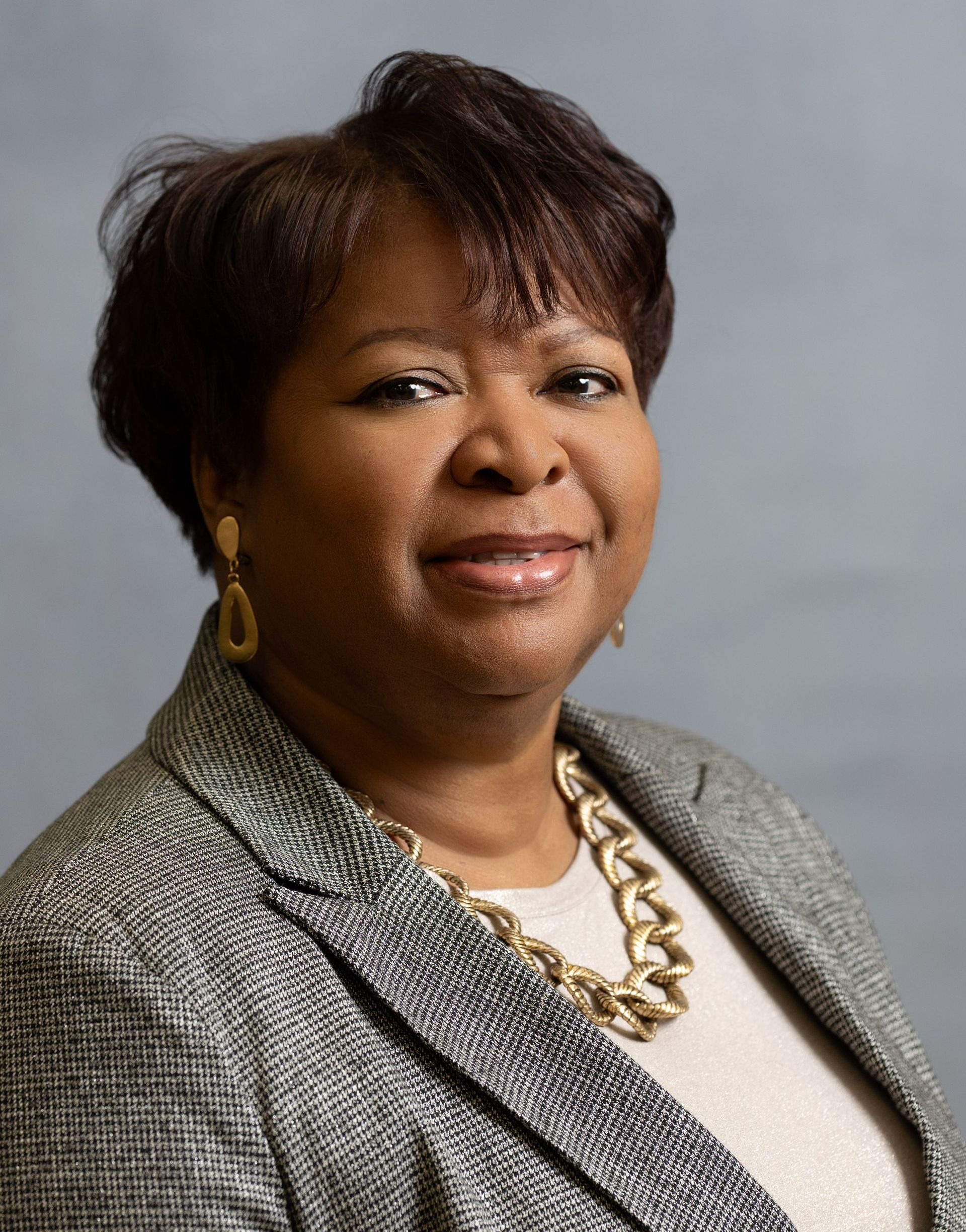 A woman with short dark hair wearing a gray patterned blazer and a chunky gold necklace against a gray background.