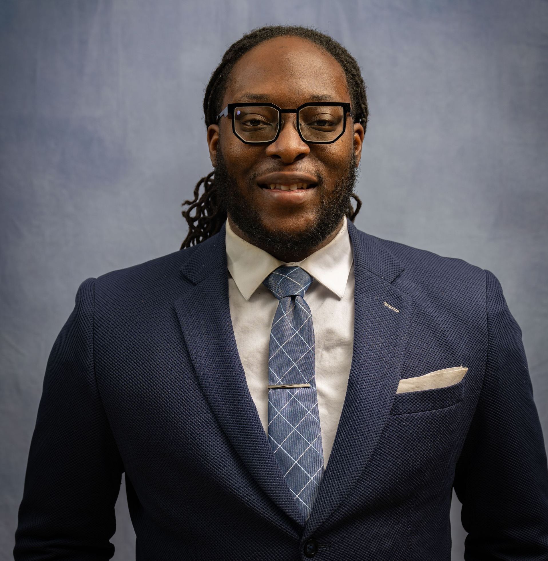A person with dark hair styled in locs, wearing glasses, a navy blazer, white shirt, and patterned tie with a tie bar.