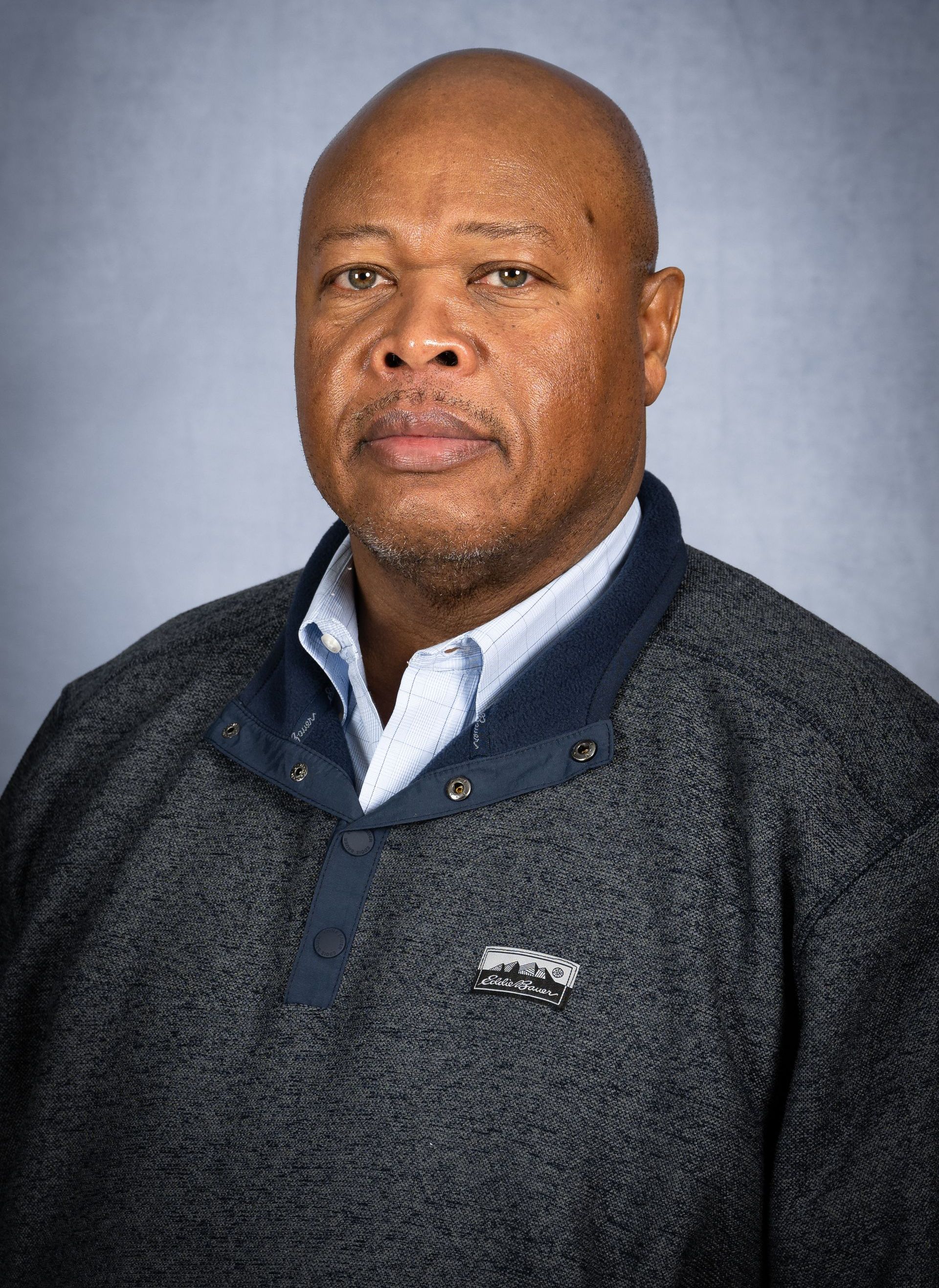 A professional headshot of a man with a neutral expression, wearing a textured grey sweater over a light collared shirt.