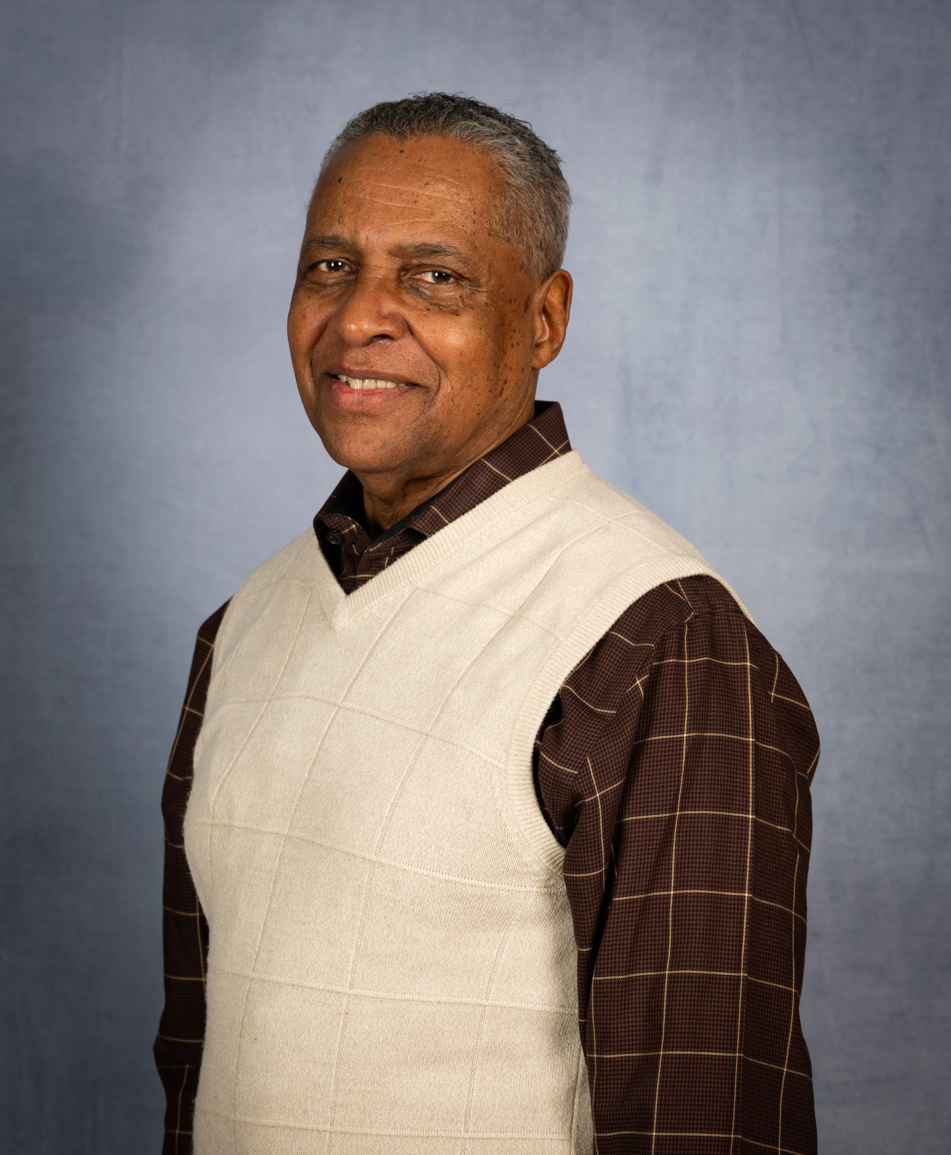 Smiling individual wearing a textured beige sweater vest over a dark brown patterned shirt against a blue background.