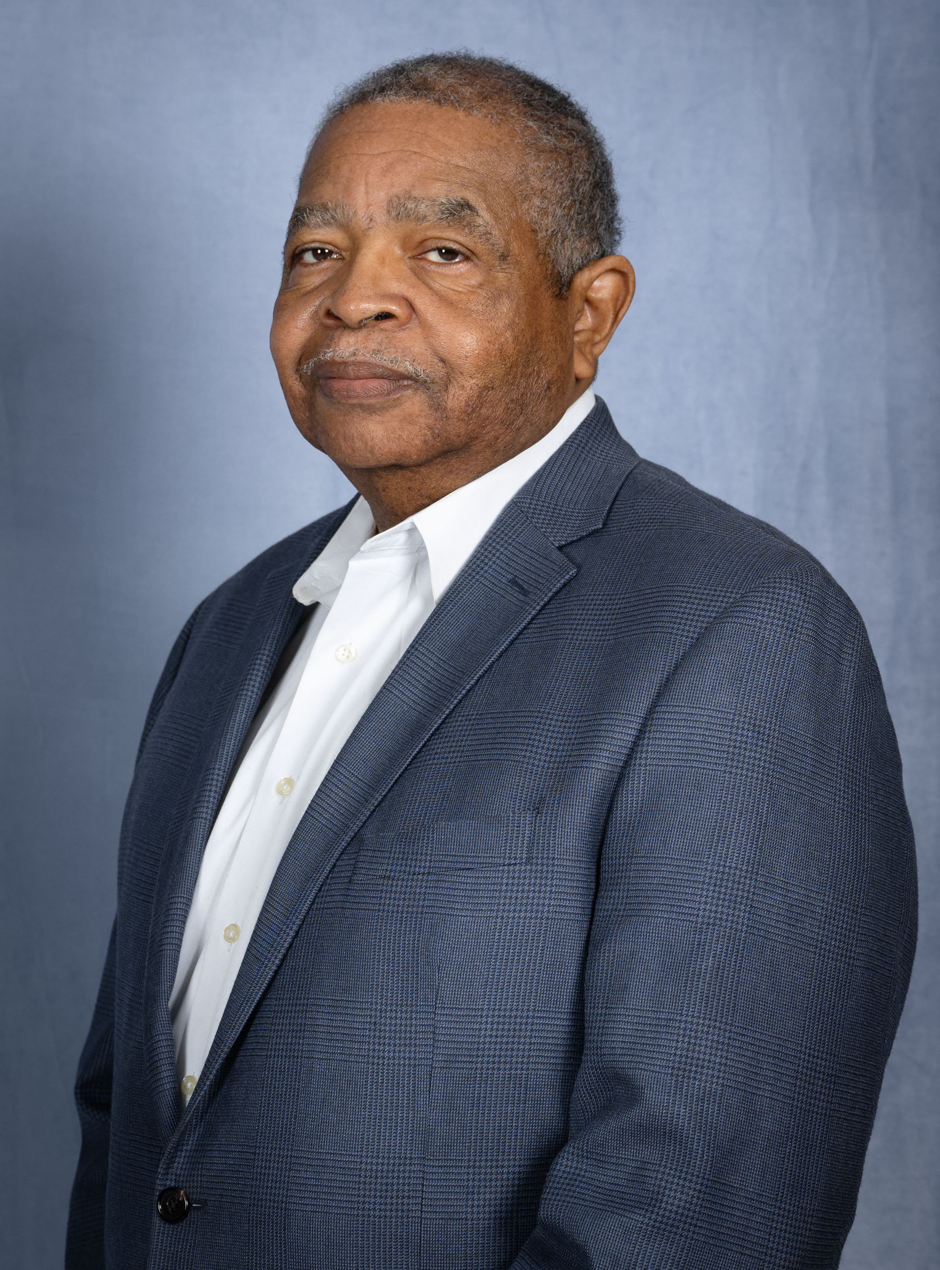 Headshot of a man wearing a white shirt and a textured blue blazer against a light blue backdrop.