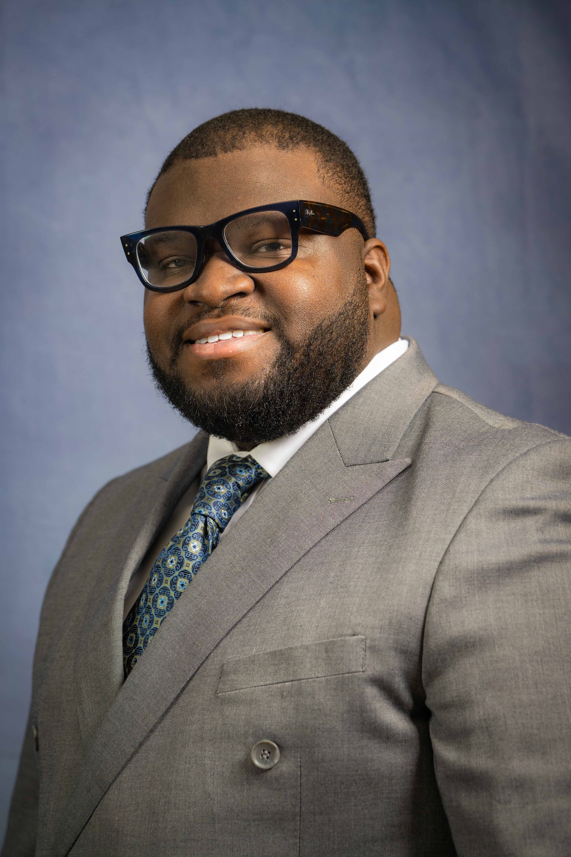 A smiling man with a beard and dark-rimmed glasses, wearing a gray blazer, white shirt, and patterned tie against a blue.