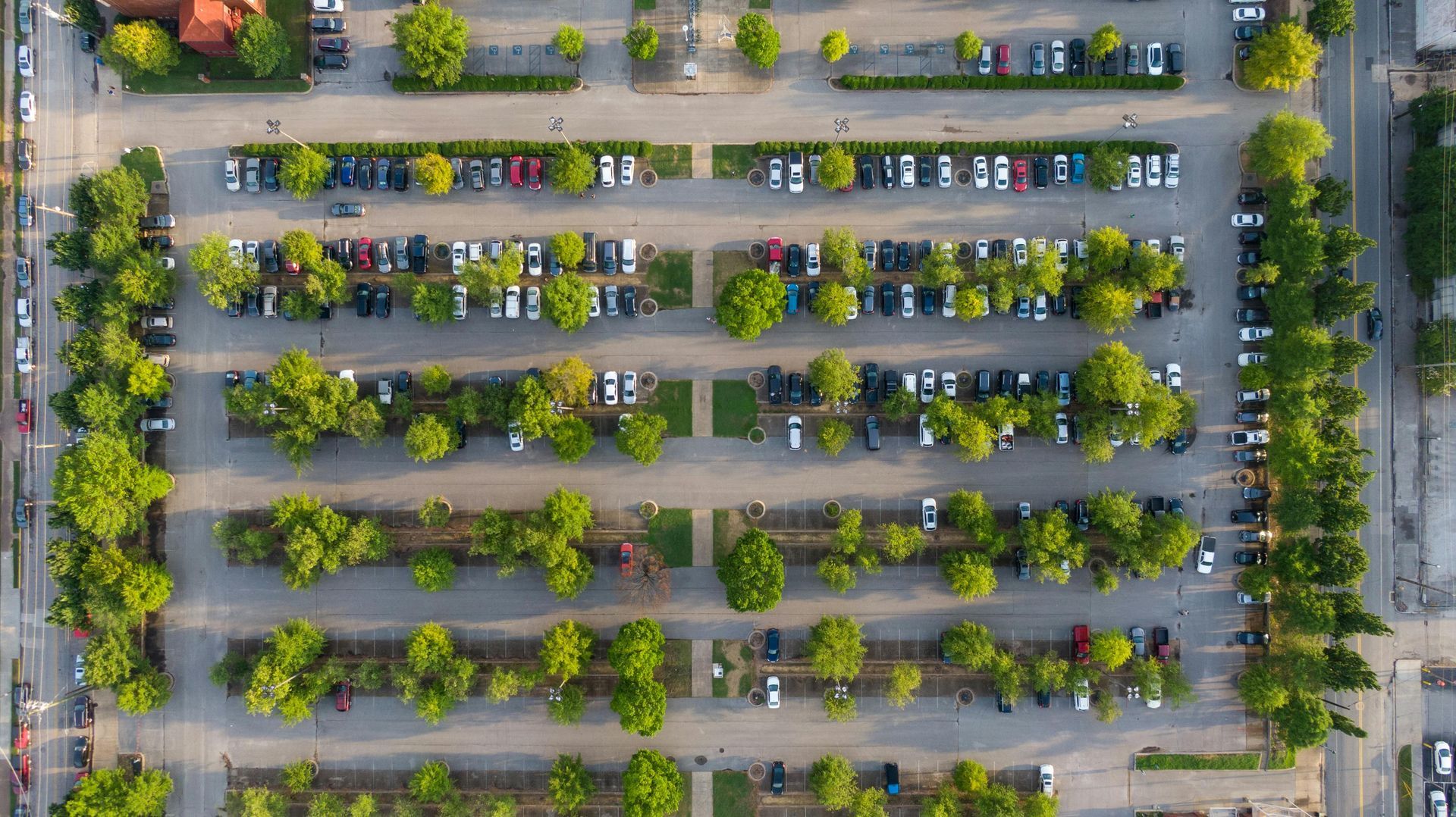 An aerial view of a large, rectangular parking lot with rows of parked cars interspersed with green trees.