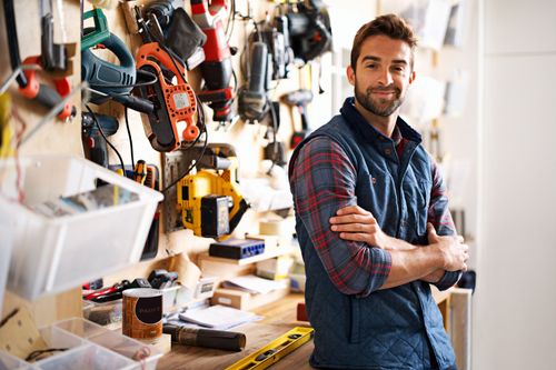 Man smiling with arms crossed in a cluttered workshop, surrounded by tools and equipment