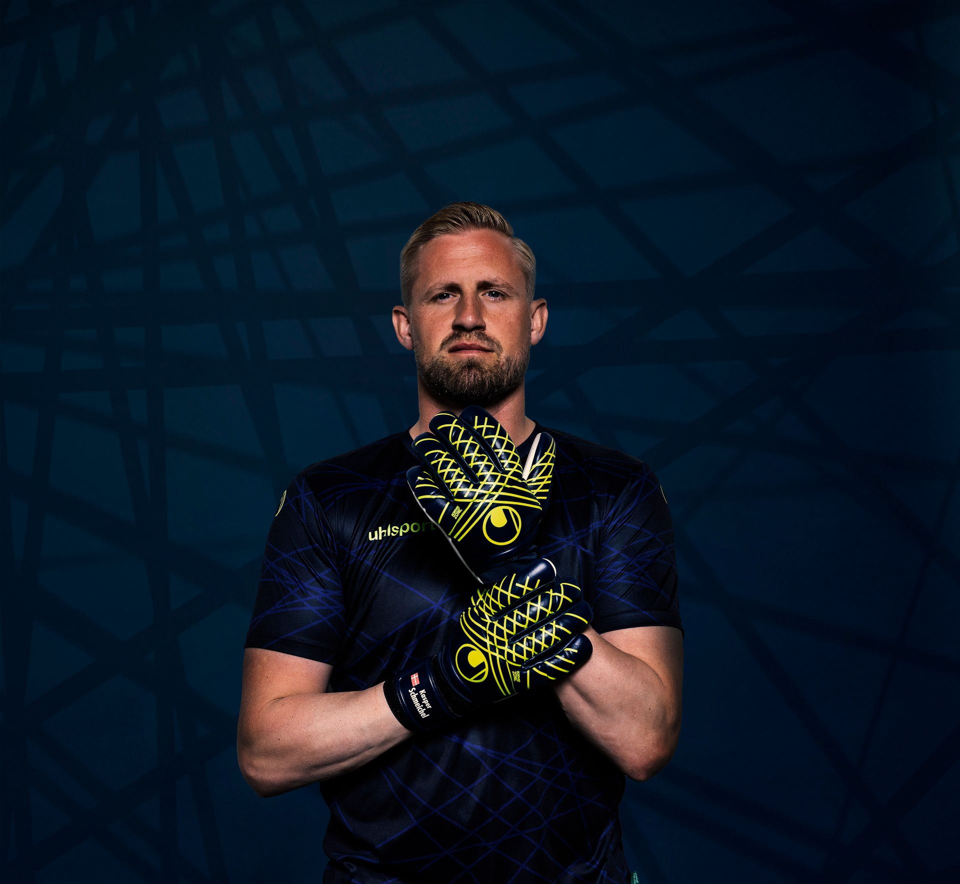 Markvörður í hönskum, blári skyrtu; með krosslagða hendur, snýr fram á við. Dökkur, mynstraður bakgrunnur.