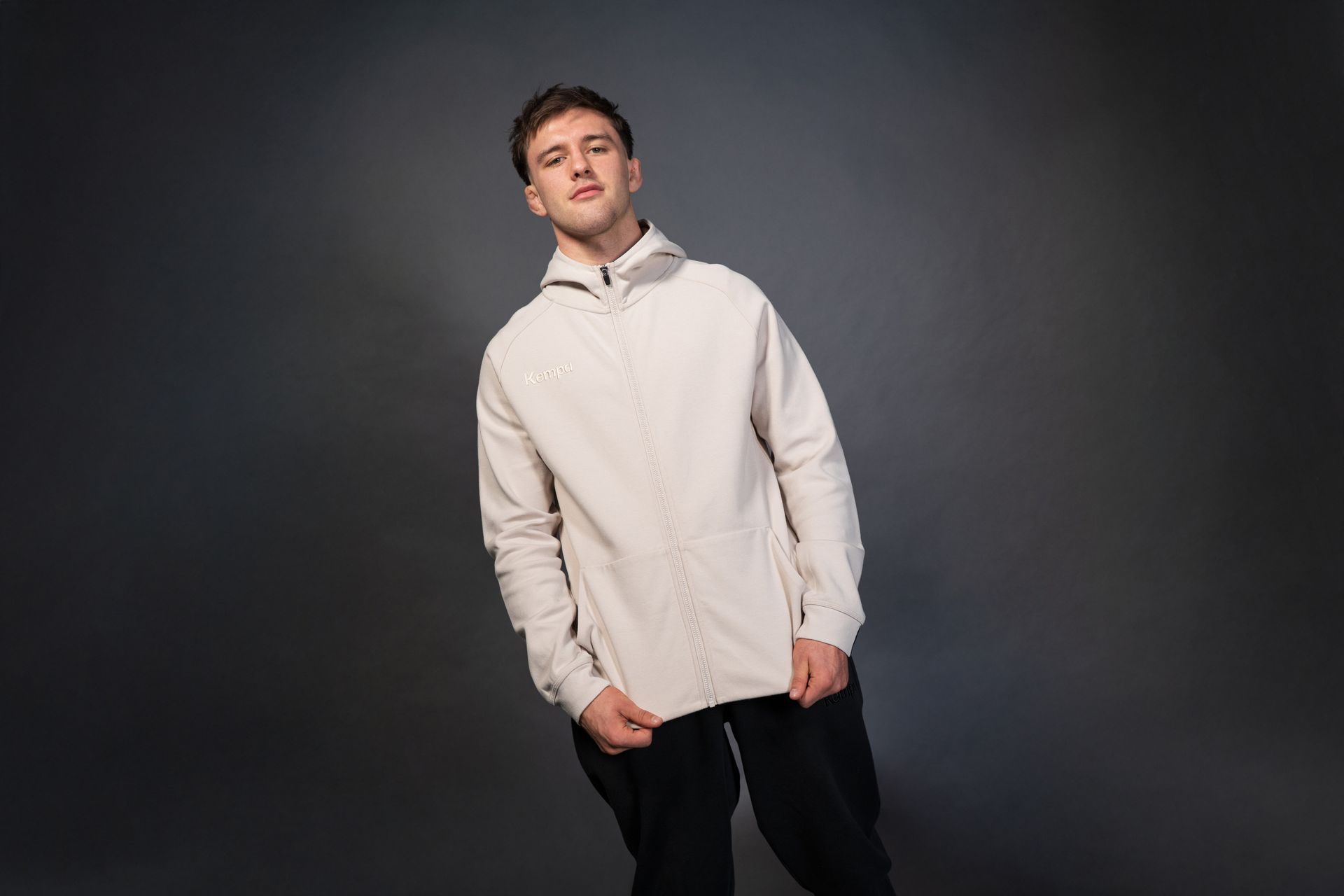 Maður í kremlituðum hettupeysu með rennilás og svörtum buxum á gráum bakgrunni, hallar sér örlítið.