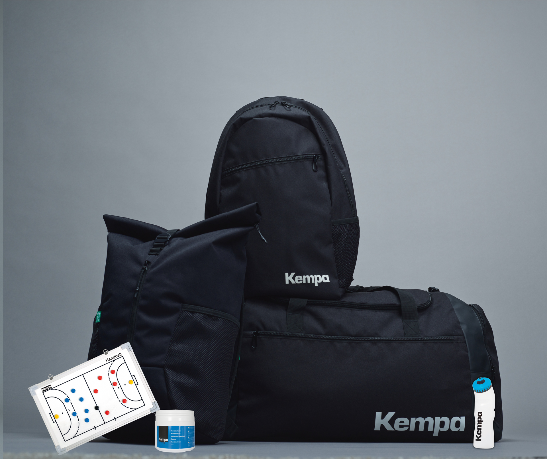 Svart íþróttatöskusett með bakpoka, vatnsflösku, hvítum töflu og rjómalit á gráum bakgrunni.