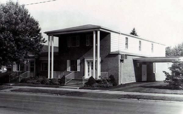 Black-and-white photo of a two-story house with a front porch, steps, and driveway beside a street