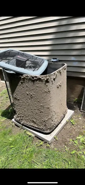 Heavily caked with dirt, an outdoor AC unit sits on a concrete pad near a house with light siding and green grass.