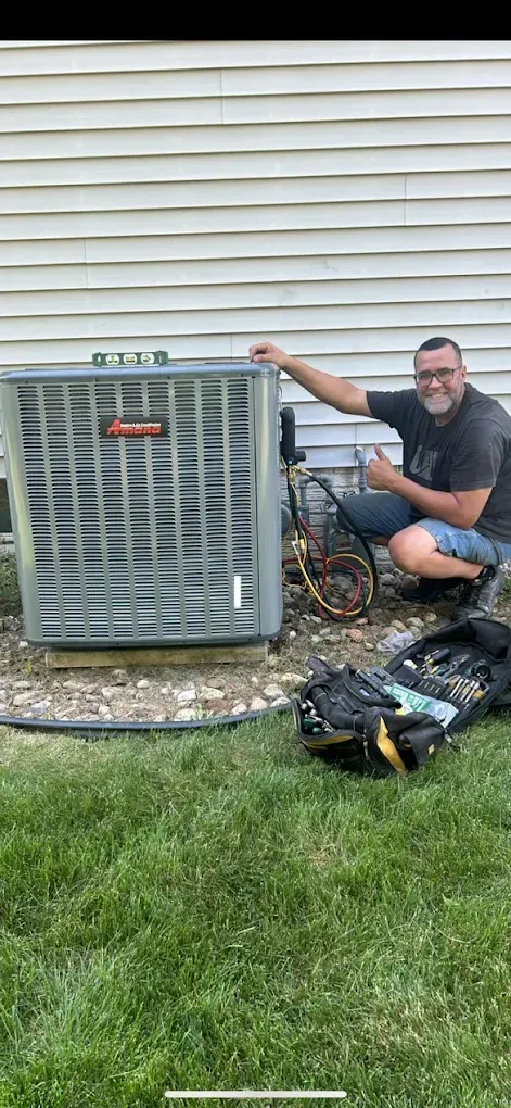 A man kneels next to an air conditioner unit, giving a thumbs up. The setting is outdoors, near a house.