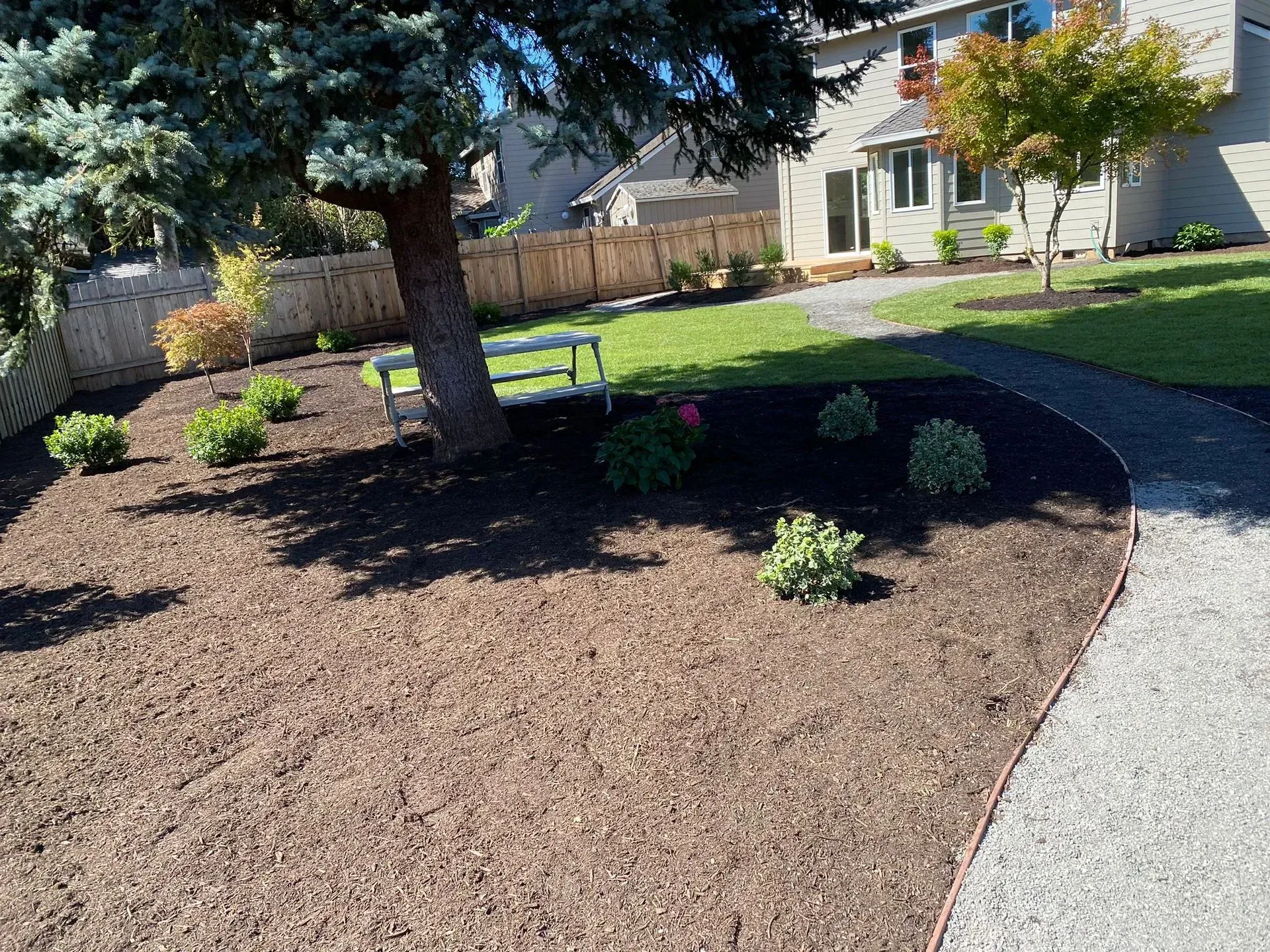 Landscaped yard with bark, plants, bench, and tree. Gravel path leads to a house.