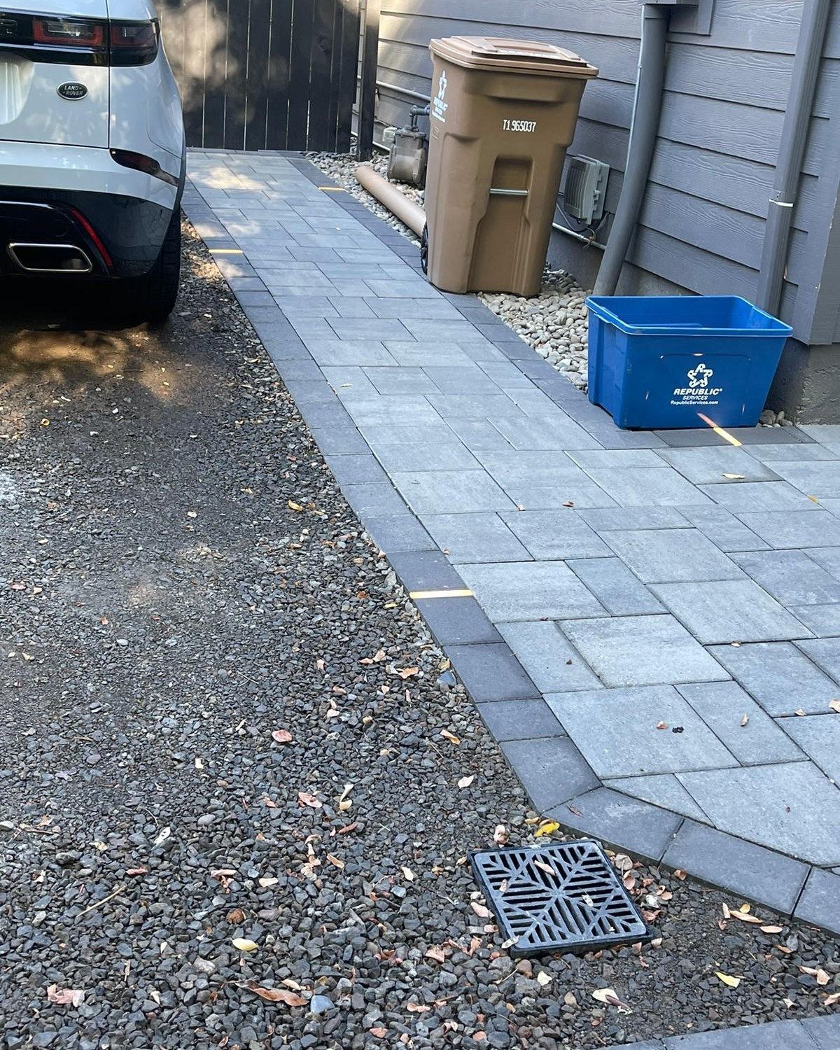 A paved pathway bordered by gravel, next to a car, trash cans, and a house exterior.