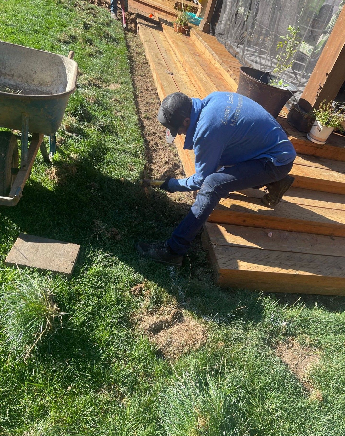 Person works on edge of wooden deck next to grass, with wheelbarrow in the background.