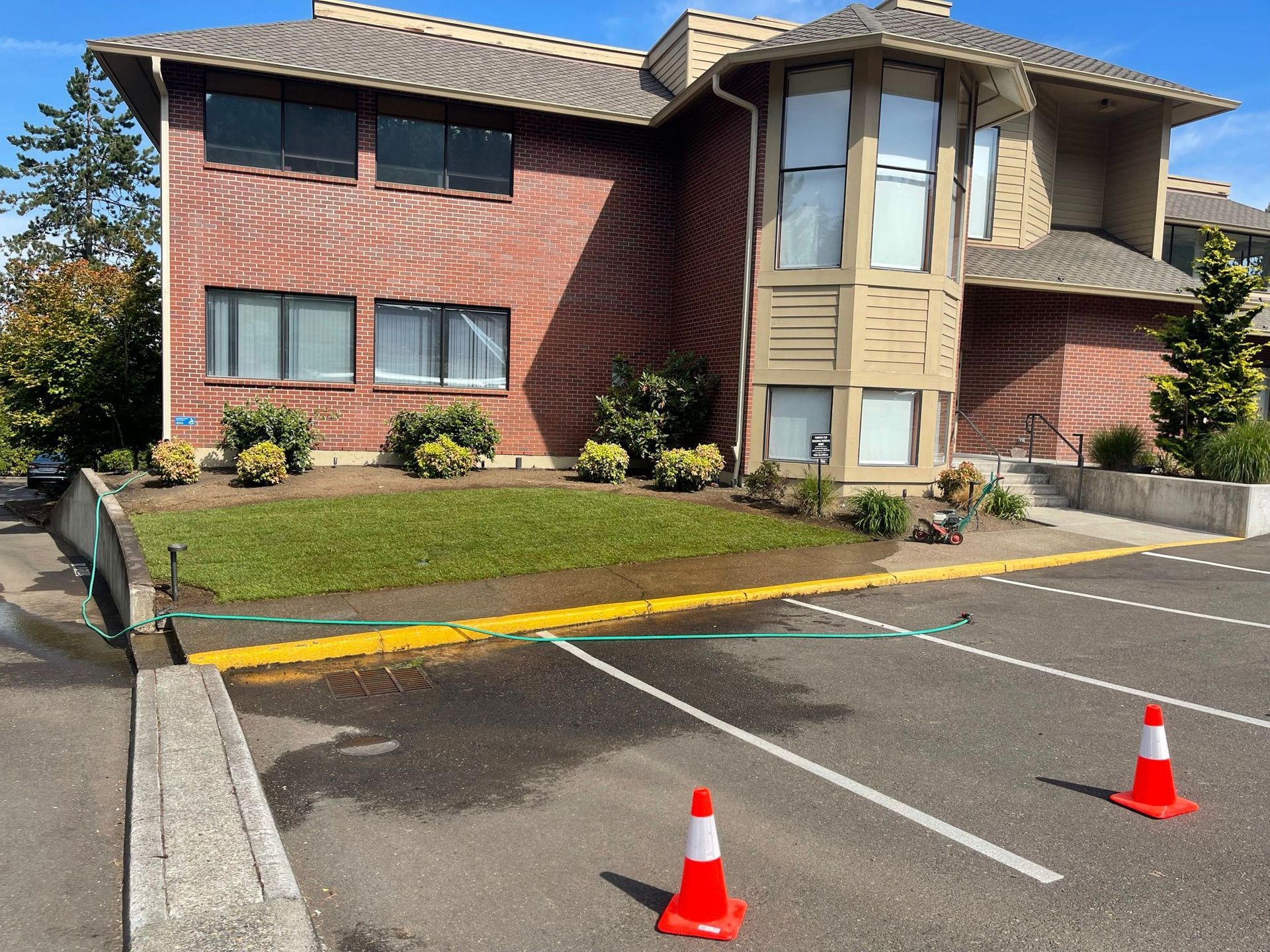 Apartment building with brick facade, grass, parking lot, and orange traffic cones. Water on the asphalt.