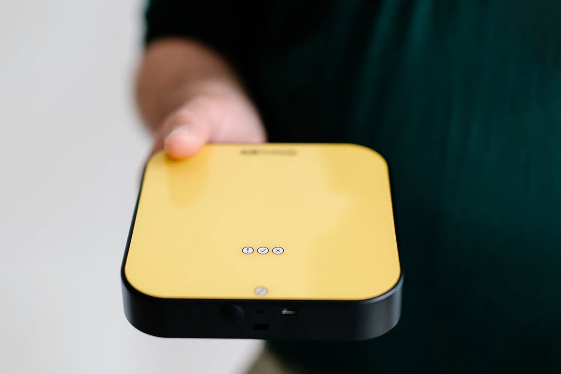 Hand holding a small yellow-and-black square device against a blurred background