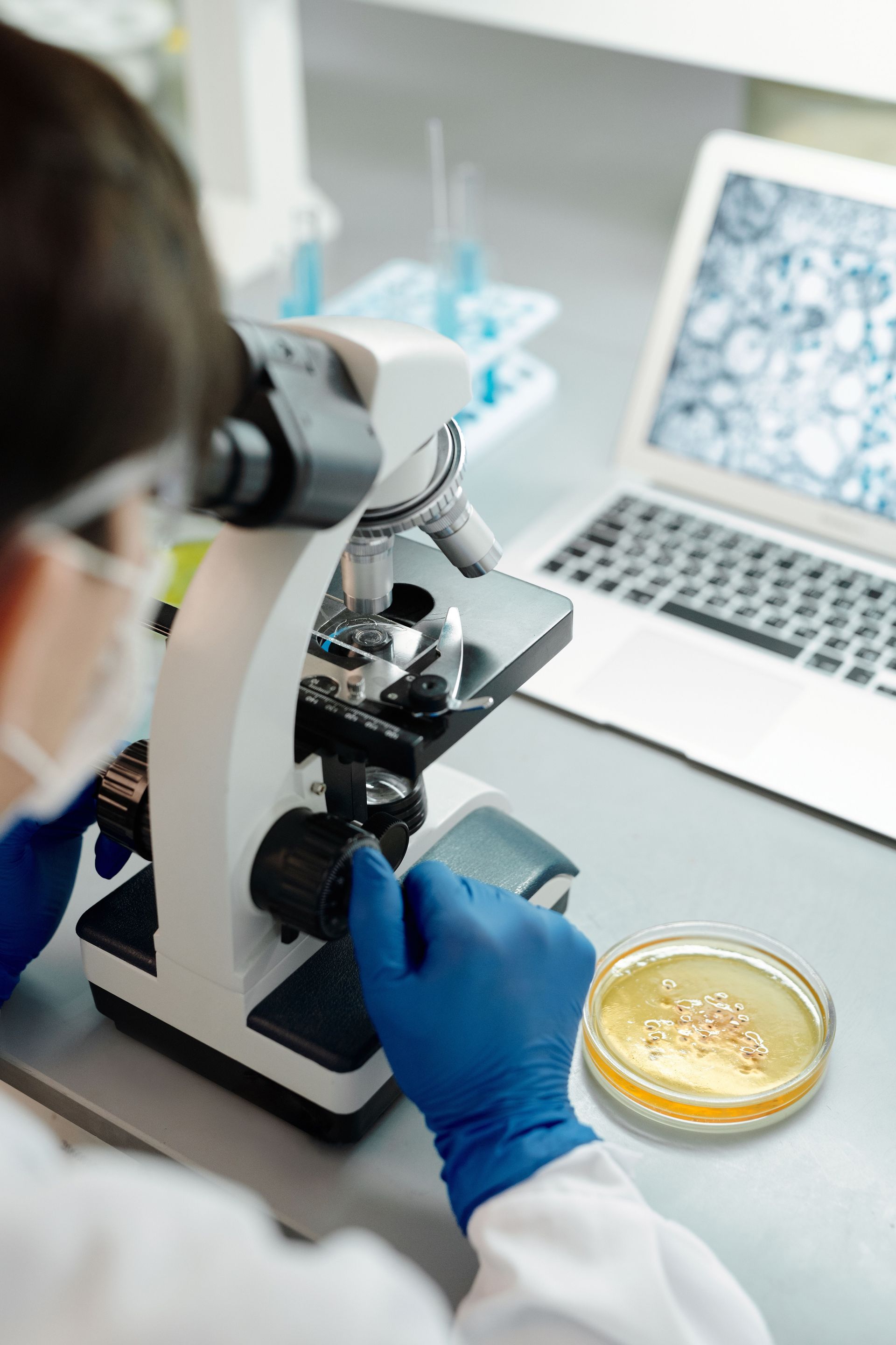 Scientist using a microscope beside a laptop and petri dish in a lab.