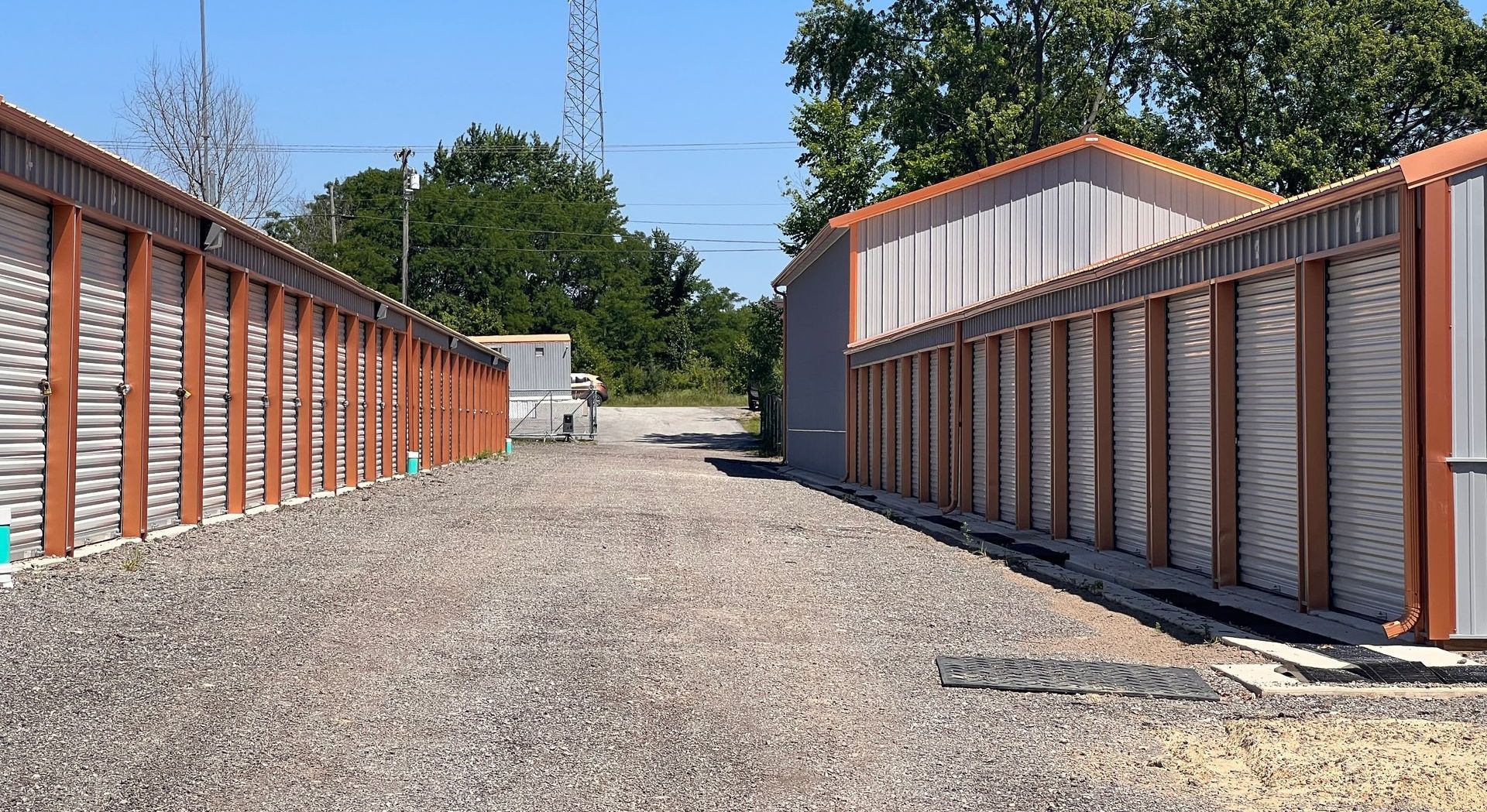 Storage units with brown frames and gray doors, gravel driveway, clear sky.