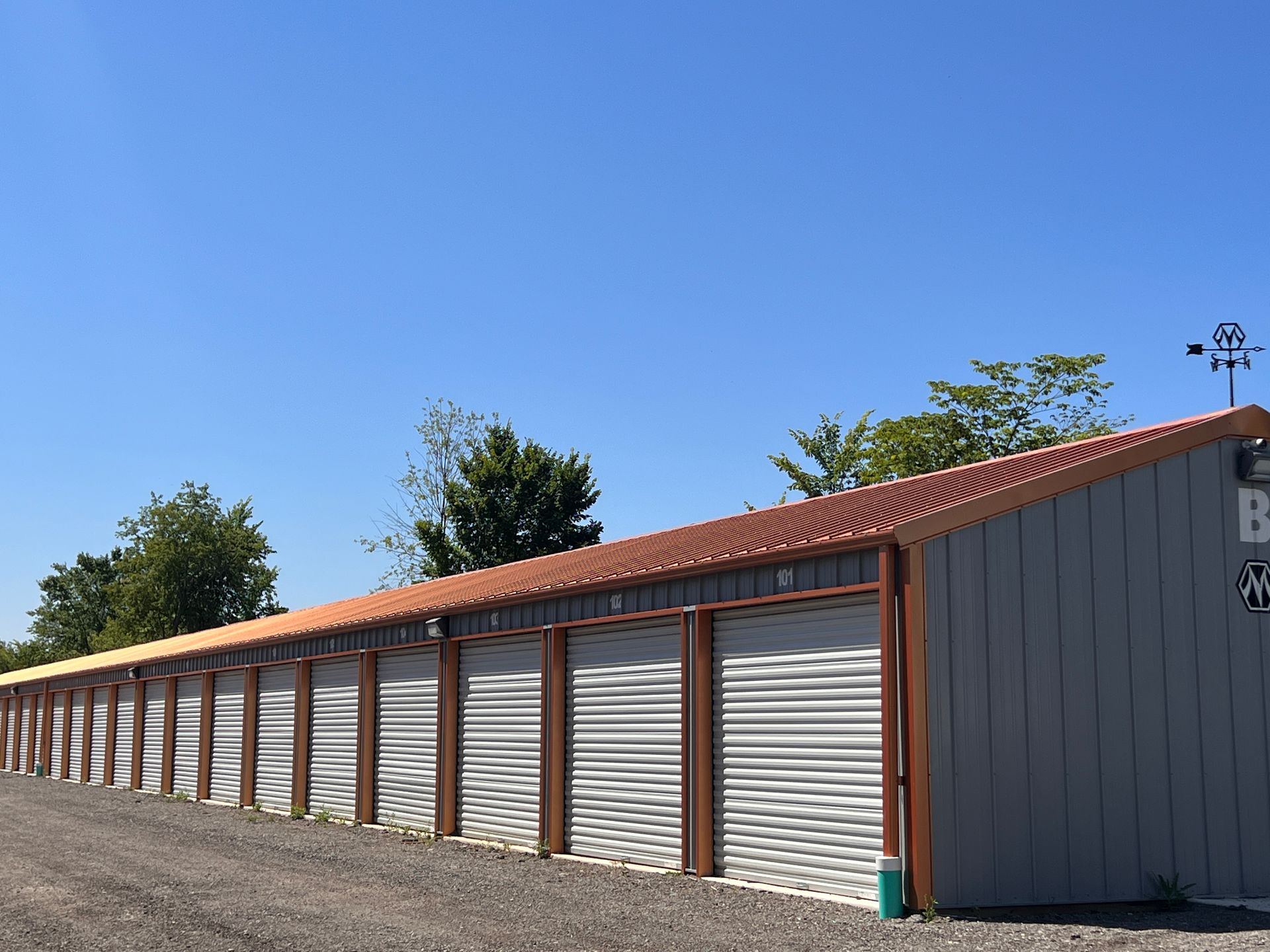 Storage units in a row, with gray doors, brown frames, and a red metal roof, under a blue sky.