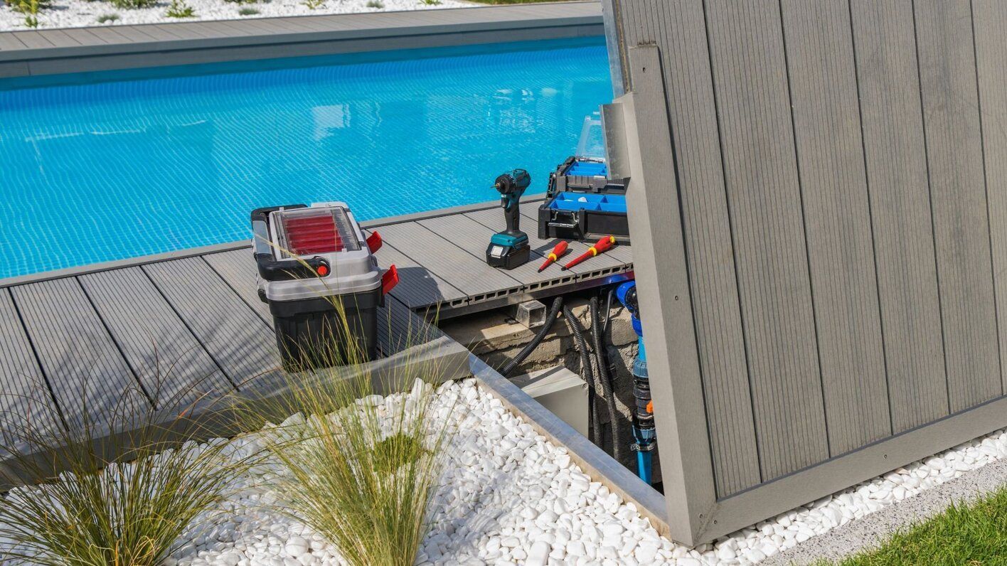 A drill and a toolbox sit on a gray pool deck next to an open access panel revealing internal machinery.