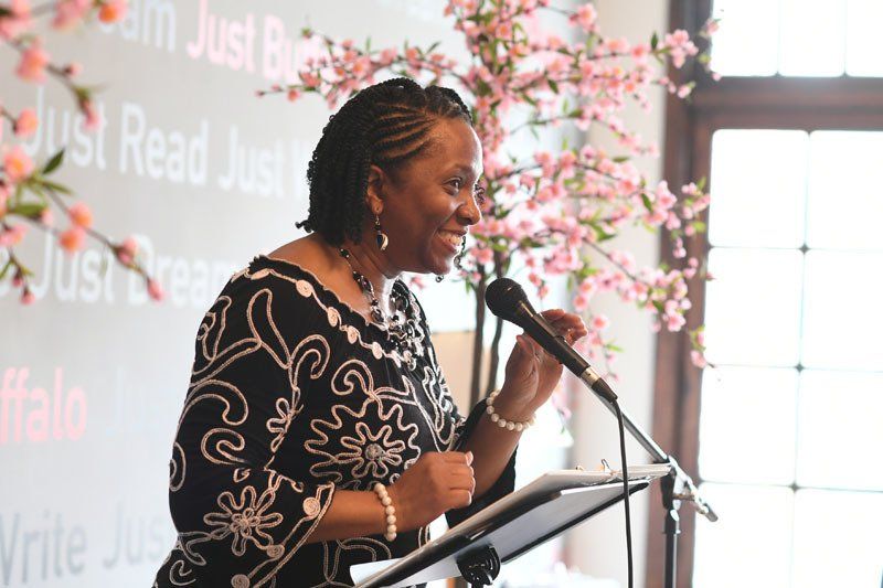 A woman is speaking into a microphone in front of a sign that says just read just write
