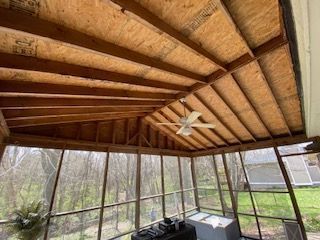 Screened porch interior with wood rafters, ceiling fan, and view of trees.