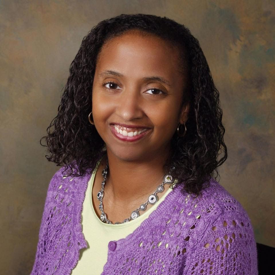 A woman wearing a purple sweater and necklace smiles for the camera